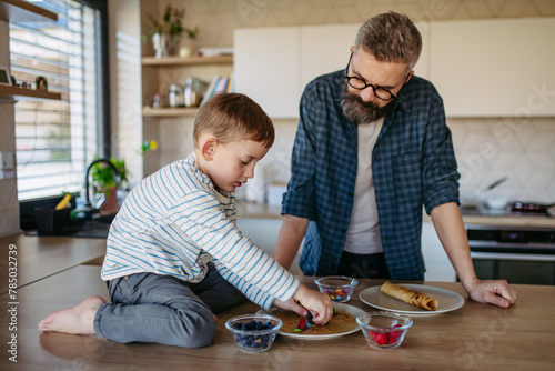 Boy filling pancake with fruit, sweets. Father spending time with son at home, making snack together, cooking. Fathers day concept. photo
