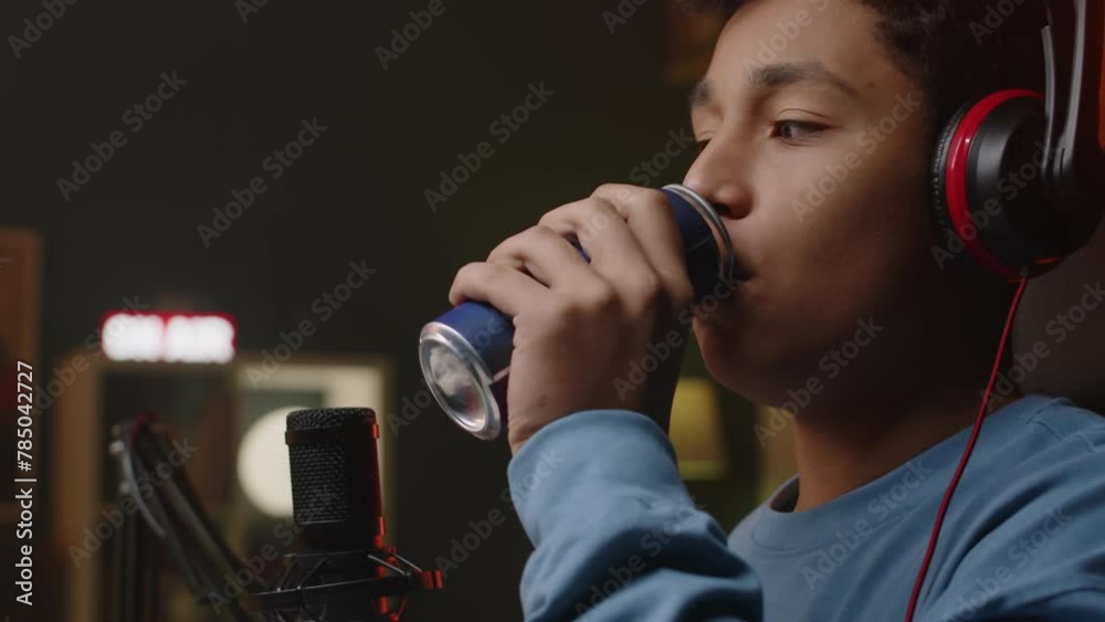 Tilting up shot of young Biracial male gamer opening can with energy drink and taking sip while doing live game streaming and playing at neon light room at night