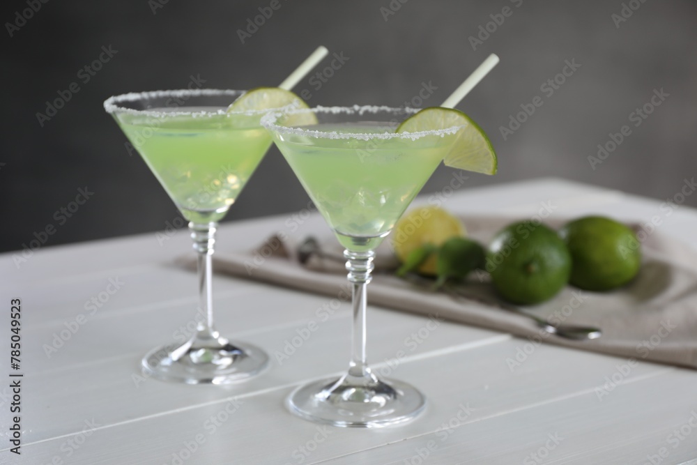 Delicious Margarita cocktail in glasses, lime and bar spoon on white wooden table