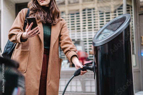 Businesswoman with smart phone holding electric plug at vehicle charging station