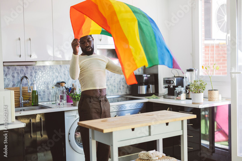 Young non-binary person holding rainbow flag near table at home photo