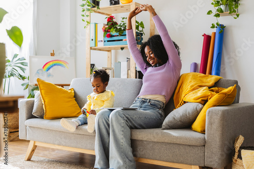 Smiling young mother stretching with hands raised sitting on sofa by daughter in living room photo