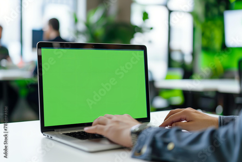  Laptop with green screen mockup on office desk, a person working in the background.