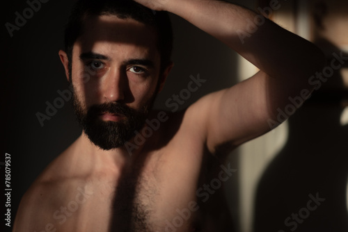 Shirtless bearded man with arm raised, looking directly at the camera. Lighting from the window sets a dramatic tone, with a heavy shadow across his face.