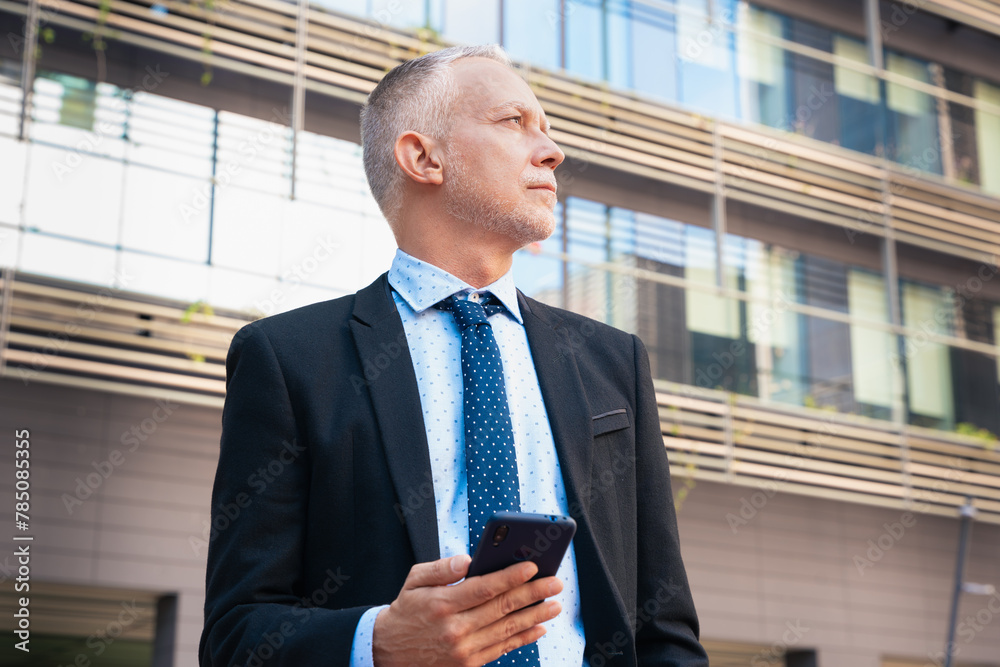 Satisfied with the results, the businessman walks down the street outside the office building, a mature boss holds a phone in his hands, writes messages and reads news online, using an app outdoors. Satisfied with the results, the businessman walks down the street outside the office building, a mature boss holds a phone in his hands, writes messages and reads news online, using an app outdoors.