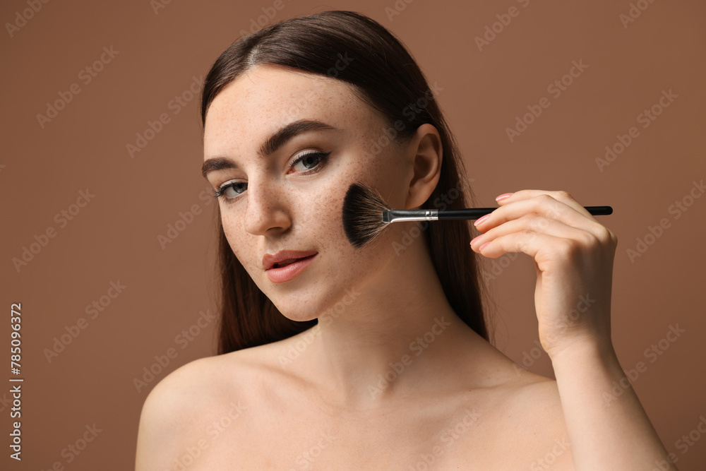 Fototapeta premium Beautiful woman with freckles applying makeup with brush on brown background