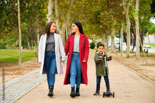 Hispanic lesbian couple walking in a park with their son in a scooter. LGBT family.