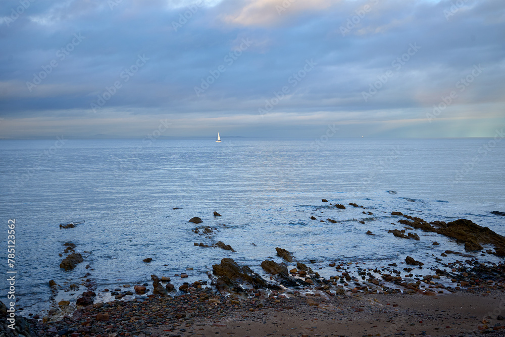 A serene coastal scene with a rocky beach, a sailboat gliding across tranquil waters, rugged terrain, and a serene sky adorned with fluffy clouds. A serene coastal scene with a rocky beach, a sailboat gliding across tranquil waters, rugged terrain, and a serene sky adorned with fluffy clouds.