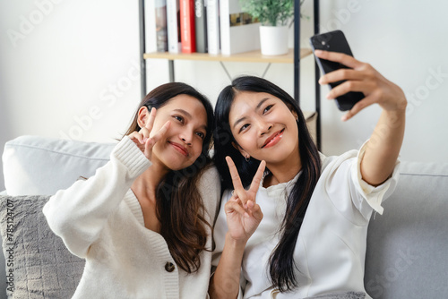 Two women are smiling and taking a selfie sitting on the sofa in the living room at home.