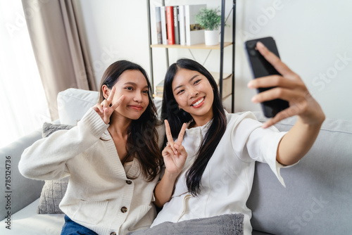 Two women are smiling and taking a selfie sitting on the sofa in the living room at home.
