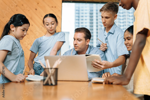 Teacher sitting and giving a lecture on lesson to children in classroom using laptop, talking to children about content in notebook. Students asking about subject instructor teach them. Edification. © Summit Art Creations