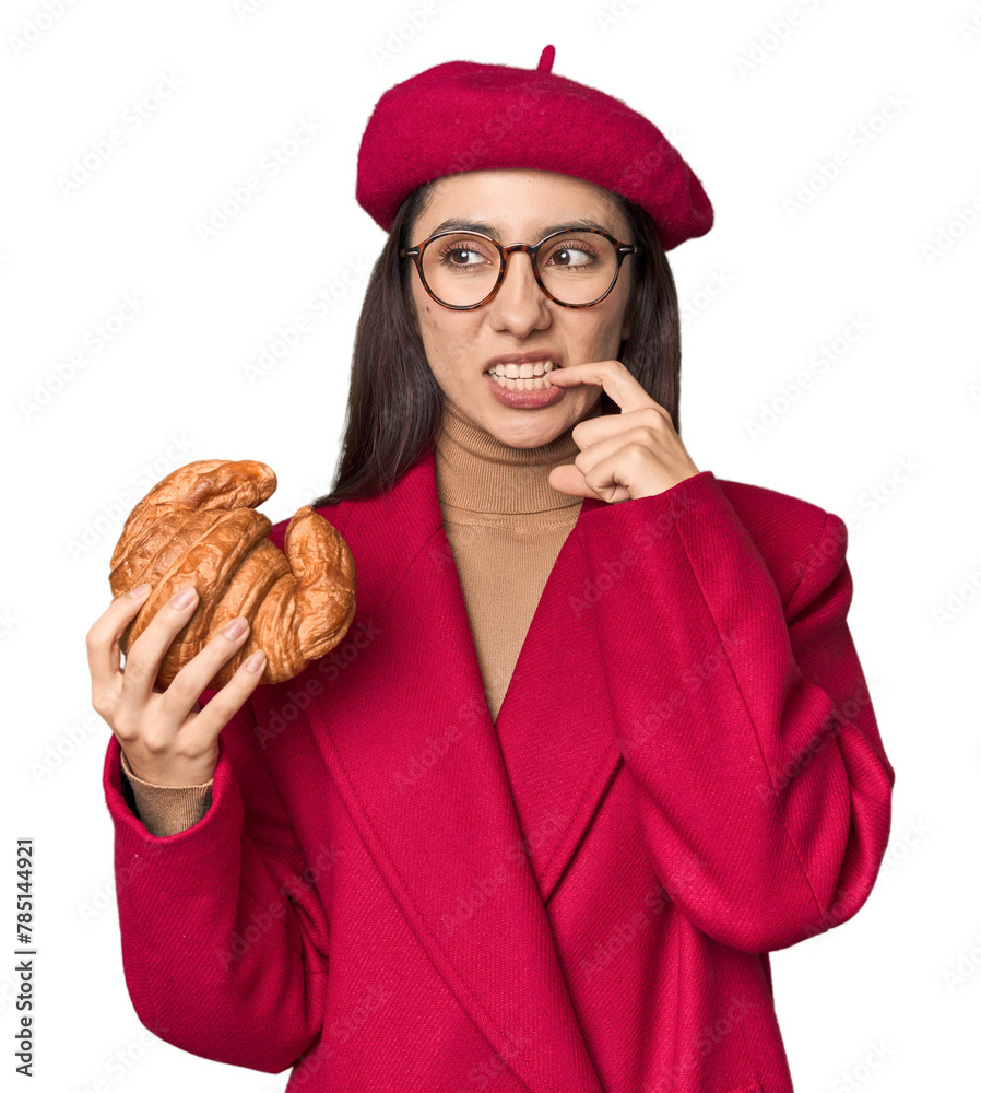 Chic Caucasian woman eating croissant in red coat and beret on studio background relaxed thinking about something looking at a copy space. Chic Caucasian woman eating croissant in red coat and beret on studio background relaxed thinking about something looking at a copy space.