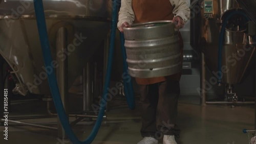 Slowmo of Biracial female worker in apron pressing buttons on control panel of stainless-steel beer tank, lifting heavy beer keg and walking with it along brewery