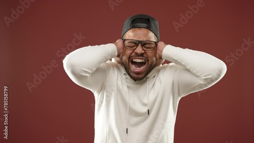 In distress, a young man with a cap and braces covers his ears with his hands, visibly pained by a loud, high decibel noise against a warm brown backdrop. High-quality 8K resolution footage.  photo