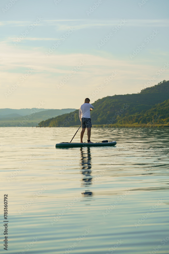 A man is standing and paddling on a paddleboard on the lake at sunset. Enjoying the vacation.  Landscape in the background. Training SUP board.