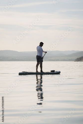 Silhouette of a man paddling on a stand up paddle boarder or SUP on the lake. Holding a paddle. Landscape in the background. Active lifestyle.