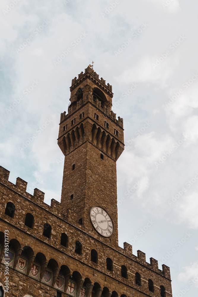 Palazzo Vecchio's imposing façade, adorned with medieval shields, from Piazza della Signoria. Torre di Arnolfo, with a clock and crowned with battlements, stands tall against a grey cloudy sky.