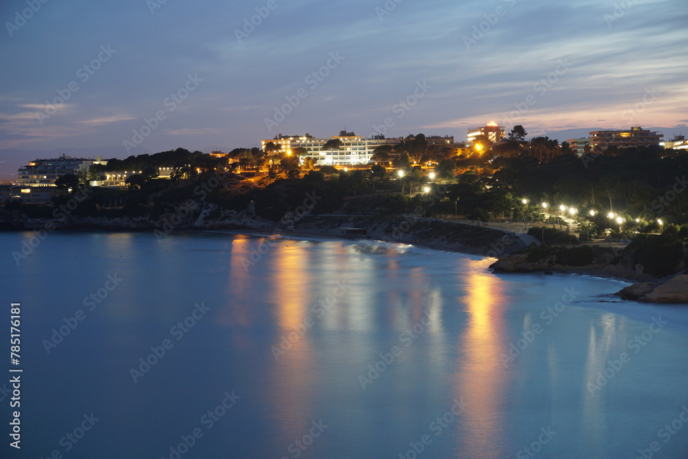 Twilight Serenity Over Mediterranean Coastal Town With Reflective Water Surface. A tranquil coastal town on the Mediterranean basks in the warm glow of twilight. Street lights begin to dot the scene.