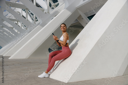 Smiling woman in sportswear sitting outdoors after training and use mobile phone while looks away