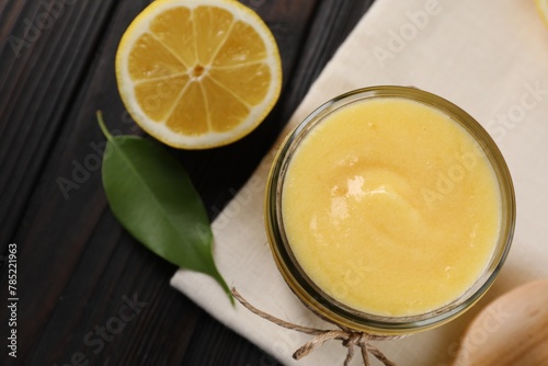 Delicious lemon curd in glass jar, fresh citrus fruit, green leaf and spoon on wooden table, top view