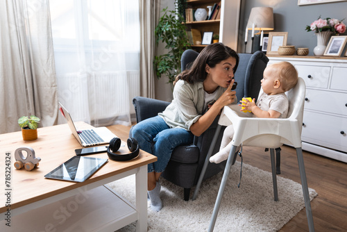Young mother, business woman preparing for work from home while taking care of her baby sitting in tall baby chair. Female business owner on maternity leave must work online from home.