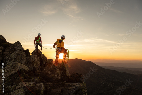Two people are climbing a mountain and one of them is holding a rope. The sun is setting in the background