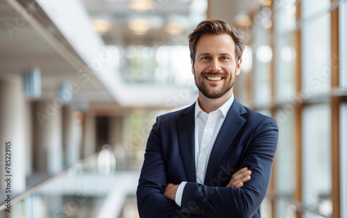 Atractivo y joven hombre de negocios con traje azul marino, brazos cruzados, con fondo desenfocado de instalaciones empresariales modernas y luminosas