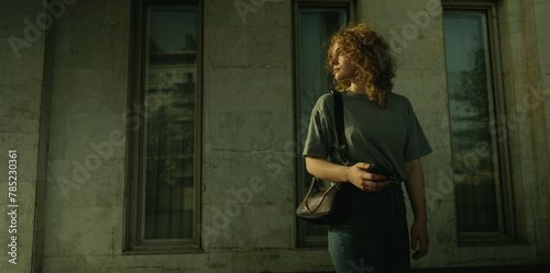 Portrait of a young curly haired woman looking back over her shoulder while holding her smart phone in her hands and standing outside in front of a city