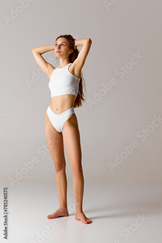 Body-size portrait of young, fit woman posing in white lingerie raising arms and put them to head against white studio background. Concept of beauty, health, depilation, anti-cellulite program, diet