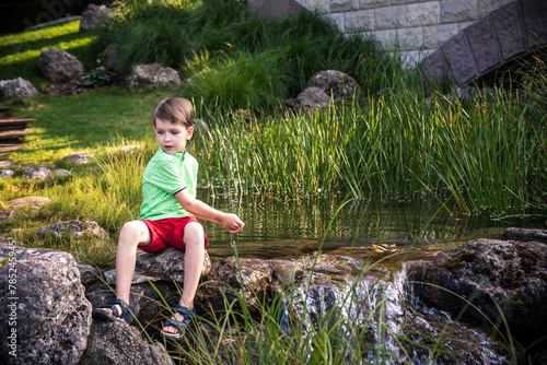 A young boy sits on the river bank, dreams. Warm summer or spring day. Cute Caucasian kid play with water