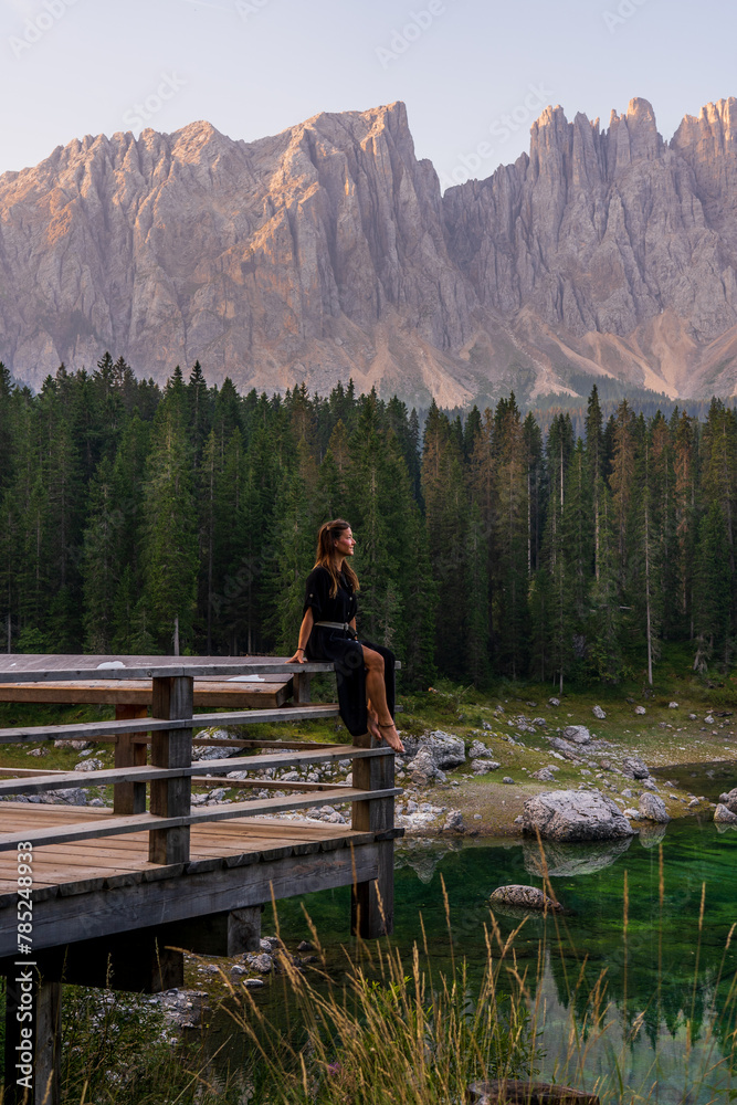 White woman in black dress sitting on the shores of an alpine lake surrounded by trees and mountains. A sense of peace and freedom. Lago Di Carezza. Dolomites. Italy