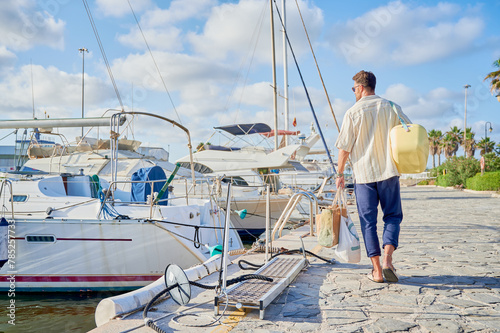 Sailing day. Handsome adult man carrying a cooling box and bags with provisions walking through to board his sailboat docked at the yacht marina.