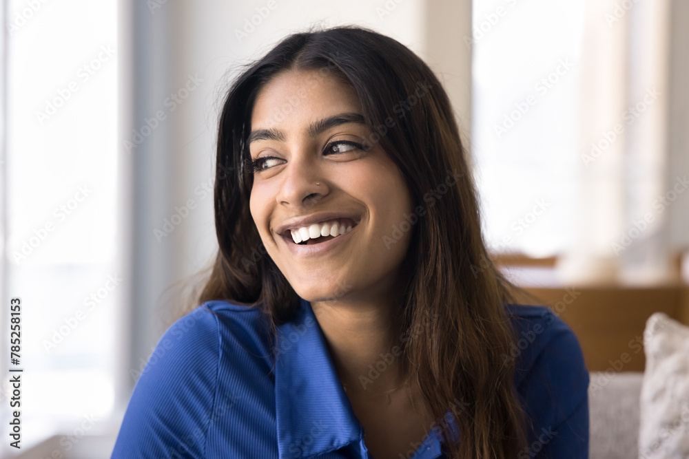Fototapeta premium Happy beautiful young Indian woman home portrait. Cheerful 20s girl with long black hair and perfect toothy smile sitting on couch, looking away in deep thoughts, thinking, smiling, dreaming, laughing