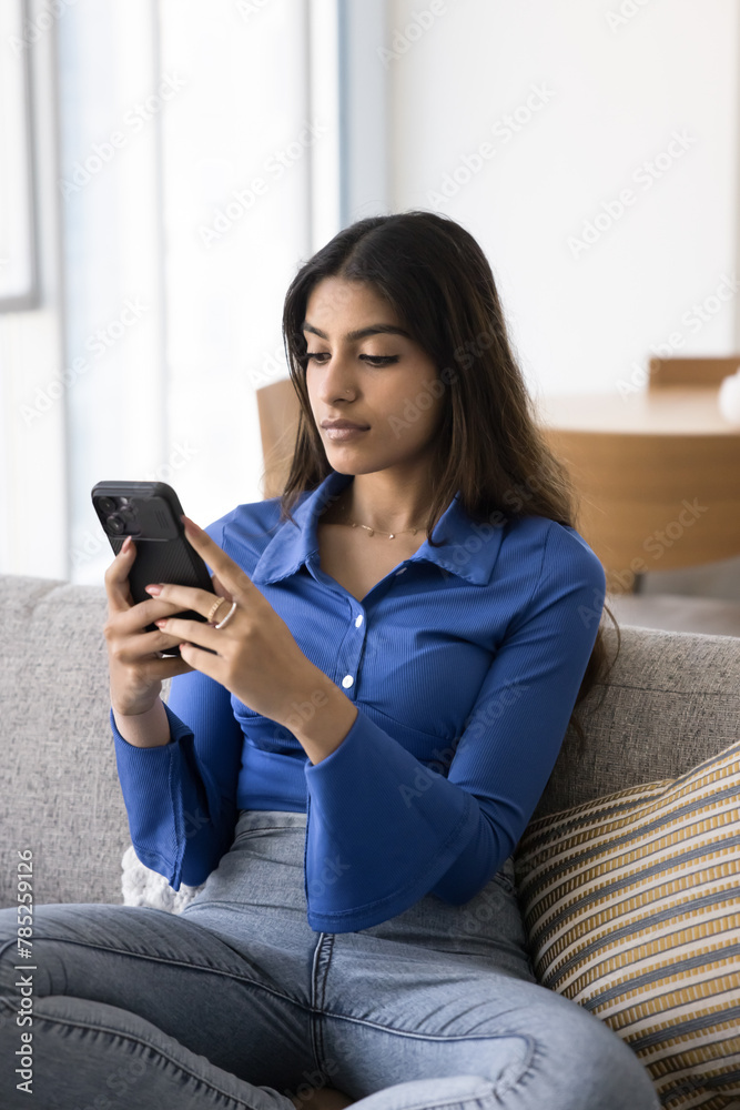 Focused beautiful young Indian woman holding smartphone, relaxing on couch at home, browsing social media, using modern wireless technology, online application, typing. Vertical shot Focused beautiful young Indian woman holding smartphone, relaxing on couch at home, browsing social media, using modern wireless technology, online application, typing. Vertical shot