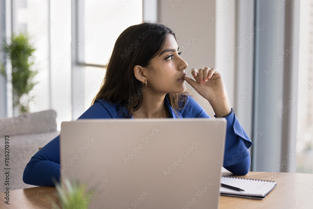 Serious thoughtful Indian freelance woman thinking on work project at laptop, looking at window away, biting thumb in deep thoughts, making decision, pondering on business plan Serious thoughtful Indian freelance woman thinking on work project at laptop, looking at window away, biting thumb in deep thoughts, making decision, pondering on business plan