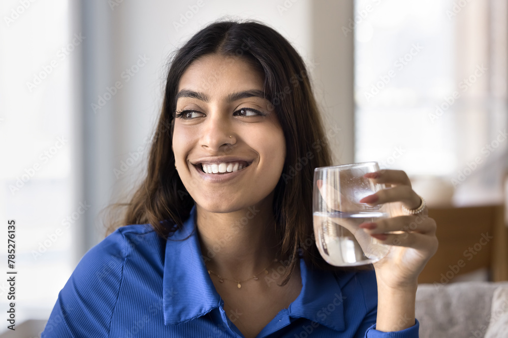 Cheerful beautiful young Indian woman promoting healthy aqua balance, holding glass of cold fresh clean water, looking away with toothy smile. 20s girl caring for healthy lifestyle, skincare