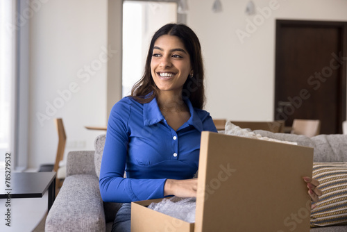 Cheerful young Indian customer girl unpacking parcel at home, holding open cardboard box, getting purchase from online store, looking away with toothy smile, laughing, thinking © fizkes