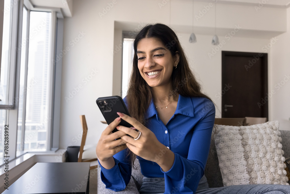 Positive excited young Indian woman typing on mobile phone, using smartphone application, online media v service for entertainment, enjoying internet communication, sitting on couch Positive excited young Indian woman typing on mobile phone, using smartphone application, online media v service for entertainment, enjoying internet communication, sitting on couch