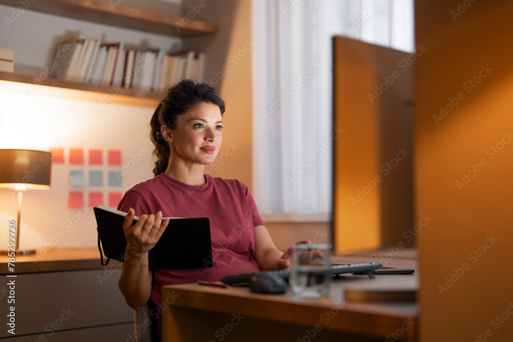 A serious mid adult woman holding her notebook while entering data on her computer A serious mid adult woman holding her notebook while entering data on her computer