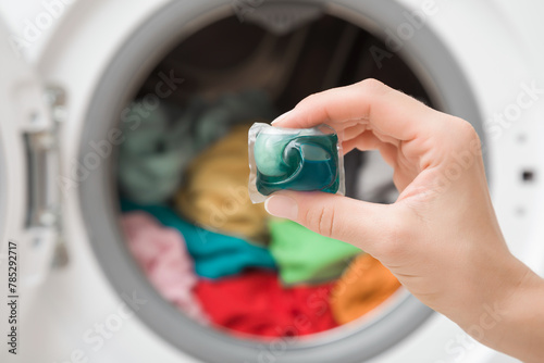 Young adult woman hand holding blue liquid capsule for colorful clothes washing on washing machine background. Closeup. Front view.