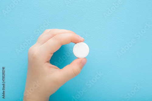 Baby hand holding white pill of c vitamin on light blue table background. Pastel color. Receiving nutrition supplement. Closeup. Top down view.