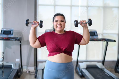 Young, Positive, Smiling, Cheerful overweight woman training with dumbbells in gym. Fat woman in sportswear doing fitness exercise. Weight loss workout, healthy lifestyle concept.