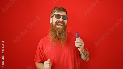 Cheerful young irish male with beard, dancing over isolated red wall, joyfully listening to funny tunes on mobile phone. handsome redhead guy smiling, holding cellphone enjoying online music.