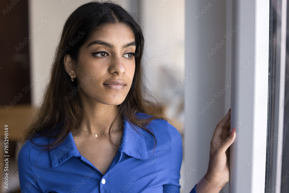 Thoughtful serious attractive young Indian woman standing at window, looking away, thinking, dreaming. Pensive beautiful girl in casual with nose stud home casual portrait Thoughtful serious attractive young Indian woman standing at window, looking away, thinking, dreaming. Pensive beautiful girl in casual with nose stud home casual portrait