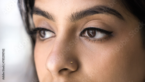 Brown eyes of young 20s Indian woman with eyeliner and mascara on extensional eyelashes. Cropped shot of face. Beauty care female model with makeup, shaped eyebrows, nose stud. Banner shot
