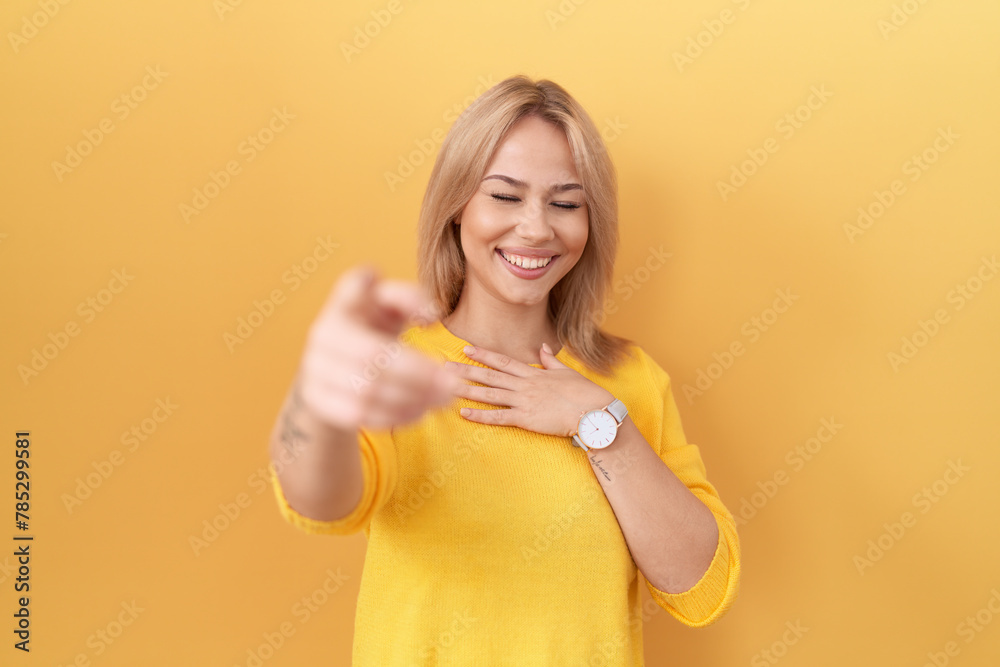 Young caucasian woman wearing yellow sweater laughing at you, pointing finger to the camera with hand over body, shame expression