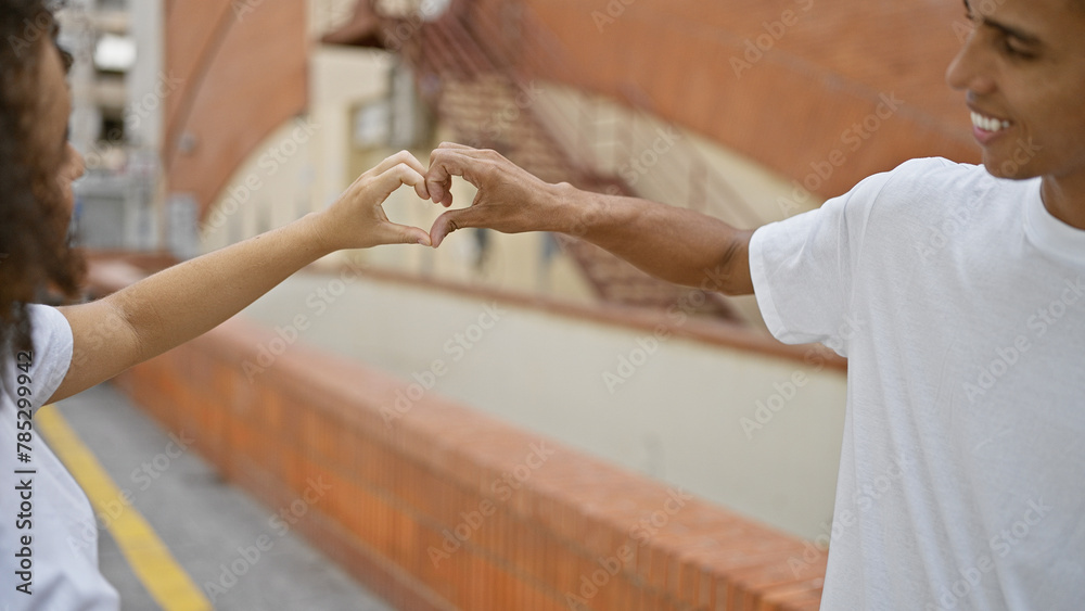 Obraz premium Beautiful couple confidently laughing & forming a heart gesture while standing united on a sunny town street, positively radiating happiness and love in their stylish, casual lifestyle.