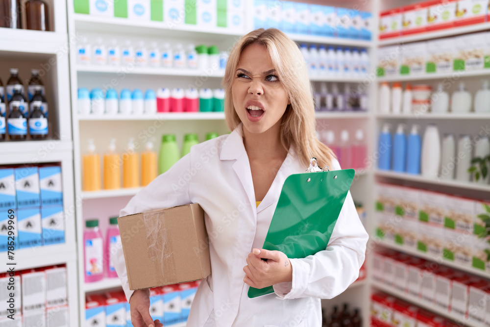 Young caucasian woman working at pharmacy drugstore holding box doing inventory angry and mad screaming frustrated and furious, shouting with anger. rage and aggressive concept.