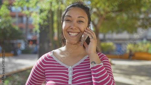A smiling young hispanic woman chats on a phone in a sunlit park, exuding joy and beauty amidst greenery and urban surroundings.