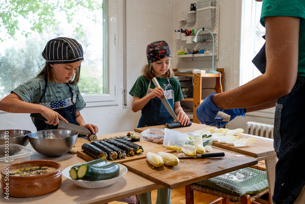 Twin sisters making maki and sushi dressed in a denim apron and cooking hat. The unrecognisable mother teaching and watching them. Twin sisters making maki and sushi dressed in a denim apron and cooking hat. The unrecognisable mother teaching and watching them.